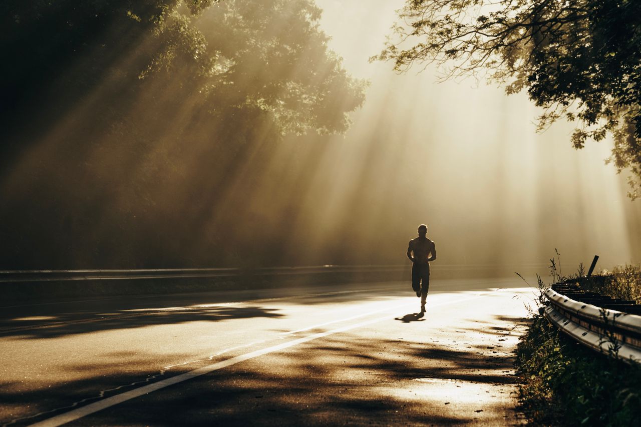 A runner jogs along a sunlit forest road, rays breaking through trees, capturing the essence of outdoor fitness.