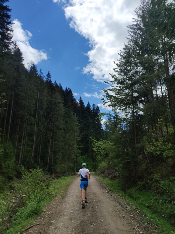 A person jogging on a forest trail with tall trees under a partly cloudy sky.